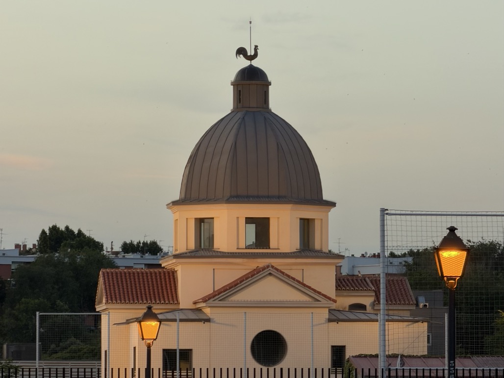 A rooster weathervane atop the slate dome of a pale classical building, glowing in warm evening light, with two lit street lamps and a sports-field fence in the foreground.