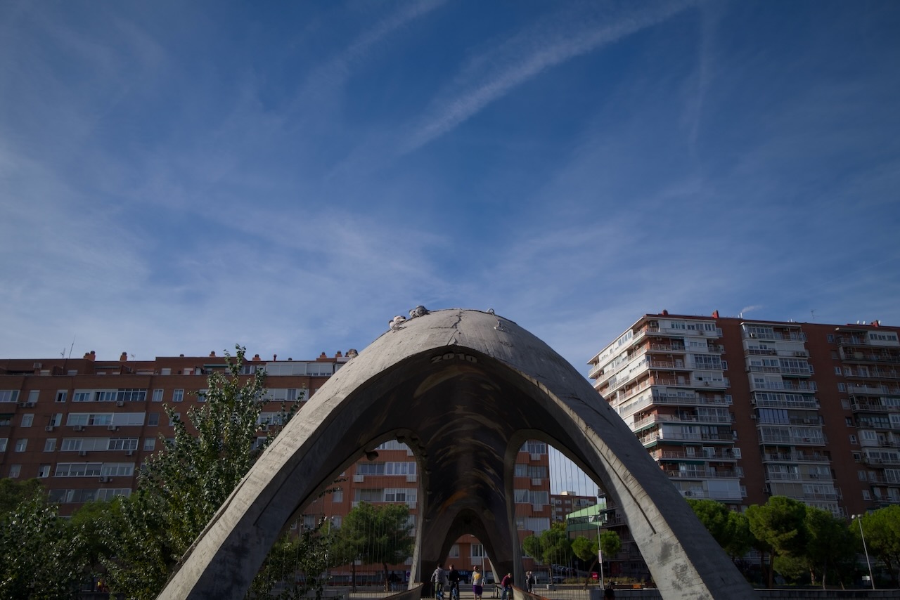 Concrete arch sculpture in front of red brick apartment buildings under a blue sky..