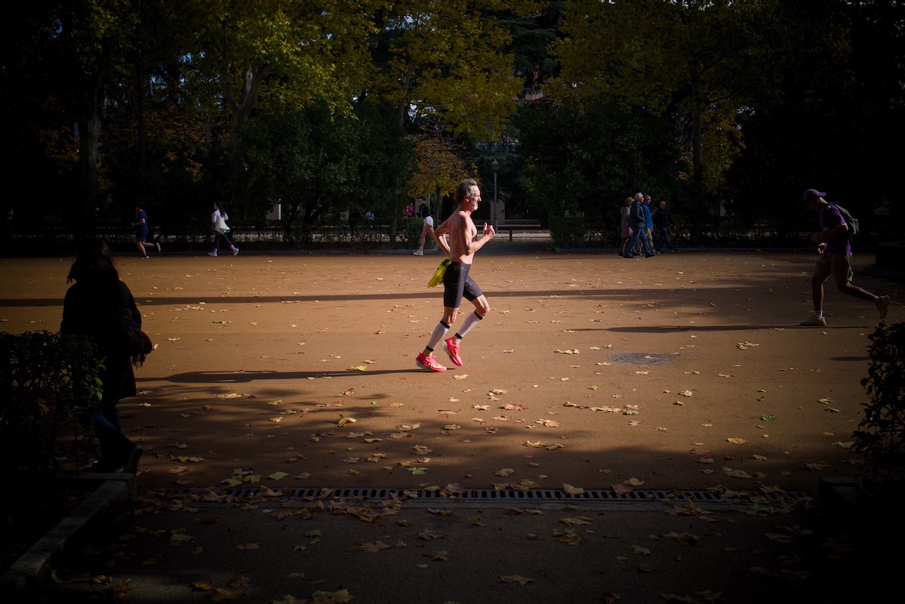 A man in red shoes jogging in a park with fallen leaves and other people around.