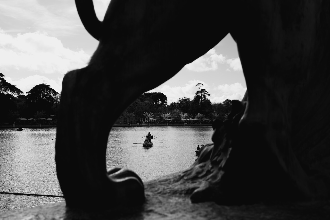 A statue frames a rower on a calm lake under a cloudy sky.