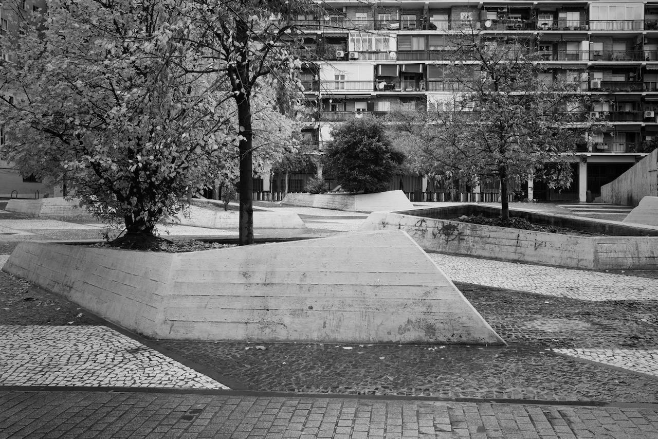 A black and white photo of a skate park with trees and buildings in the background.