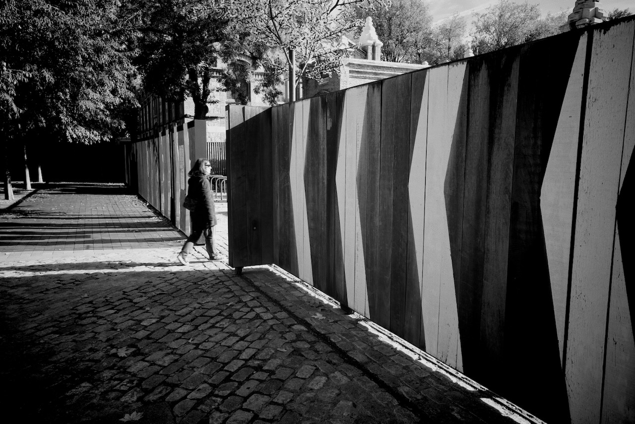 A person walks along a cobblestone path beside a tall wooden fence in a sunlit, tree-lined area.