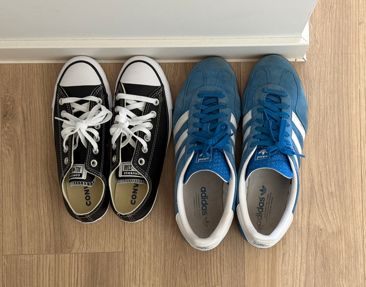 Two pairs of sneakers, black Converse and blue Adidas, placed side by side on a wooden floor.
